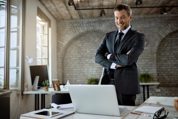 Happy male entrepreneur standing with arms crossed while reading something on a computer in the office.